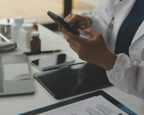 Serious female doctor using laptop and writing notes in medical journal sitting at desk. Young woman professional medic physician wearing white coat and stethoscope working on computer at workplace.